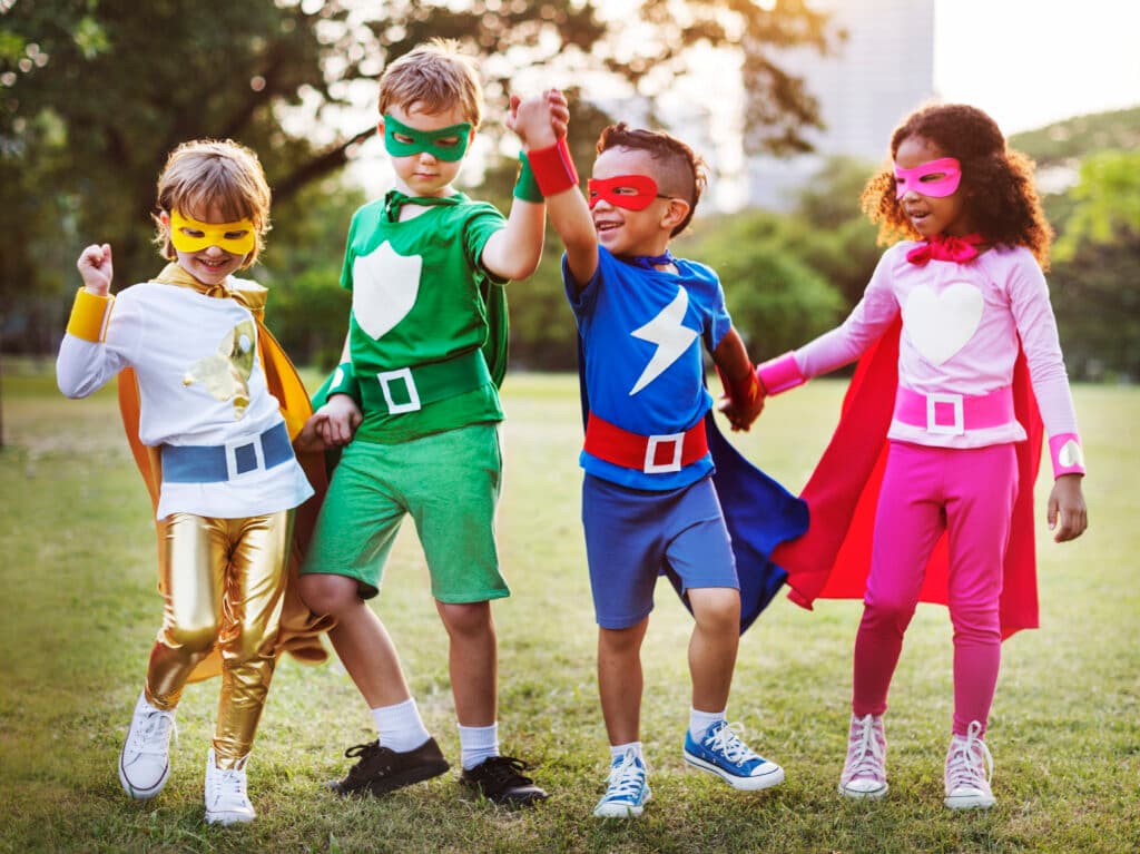 Four children in colorful superhero capes and masks playing together in a sunny park, one raising a fist to the sky